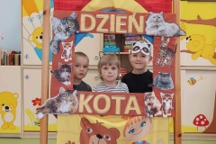 Jedna dziewczynka i dwóch chłopców stoją za przedszkolnym teatrzykiem w prostokątnej brązowej ramce. Brzegi ramki ozdobione zdjęciami kotów z literami układającymi się w napis Dzień Kota. W tle przedszkolna szafka z zabawkami i książeczkami dla dzieci.