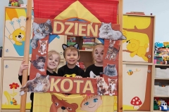 Dwie dziewczynki i jeden chłopiec stoją za przedszkolnym teatrzykiem w prostokątnej brązowej ramce. Brzegi ramki ozdobione zdjęciami kotów z literami układającymi się w napis Dzień Kota. W tle przedszkolna szafka z zabawkami i książeczkami dla dzieci.