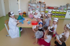 Dwie dziewczynki siedzą przy stoliku przebrane za pielęgniarki, wokół nich na podłodze siedzi reszta dzieci, na stole leżą przybory których używa pielęgniarka.