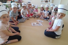 Grupa dzieci siedzi w półkolu, na głowach mają czepki jak pielęgniarki, pośrodku nich na podłodze leżą obrazki.