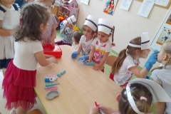 Dzieci z czepkami pielęgniarek stoją wokół stołu , omawiają przybory których używa pielęgniarka.