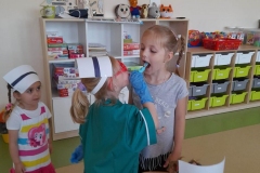 Jedna dziewczynka przebrana za pielęgniarkę bada gardło drugiej dziewczynce.