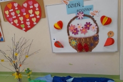 Na tablicy kosz z kolorowymi kwiatami, nad koszem napis na niebieskich kartkach"dzień rodziny". Wokół kosza upięte serduszka. Pod tablicą ułożony stawek. Obok tablicy duże, czerwone serce.
