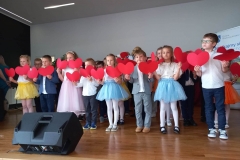 Grupa dzieci stoi na scenie, każde z nich trzyma w rękach papierowe, czerwone serce.