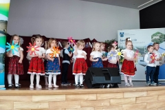 Zdjęcie przedstawia dzieci stojace na scenie. Każde trzyma w ręku kolorowe kwiaty.