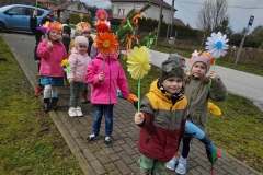 Grupa dzieci idzie parami chodnikiem, każde z nich trzyma w reku kolorowego kwiatka. W tyle dziecko trzyma wiosenny gaik.