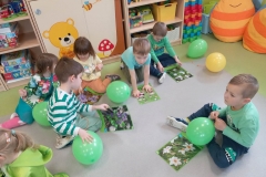 7 dzieci siedzi na podłodze,  przed nimi leżą zielone balony.