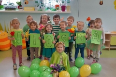 Grupa dzieci stoi w dwóch rzędach. Na zielonych kartkach widnieje napis wiosna. Na podłodze przed dziećmi leżą zielone balony