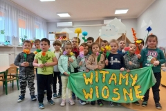 Grupa dzieci stoi w dwóch rzędach. Na zielonych kartkach widnieje napis wiosna. Na podłodze przed dziećmi leżą zielone balony.