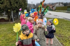Grupa dzieci idzie parami chodnikiem, każde z nich trzyma w reku kolorowego kwiatka. W tyle dziecko trzyma wiosenny gaik.