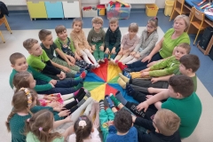 Duża grupa dzieci wraz z wychowawczynią siedzi na kolorowej chuście animacyjnej.