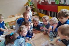 Dzieci zgromadzone są wokół stolików, nachylają się nad naczyniem z wodą w której zanurzona jest papierowa łódka. Dmuchają w nią, obserwują co się dzieje.