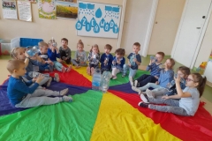 Dzieci siedzą na brzegach chusty animacyjnej, piją wodę z małych butelek. Na środku chusty widać butelki z wodą.