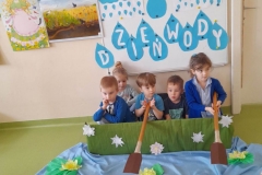 W Zielonej łódce wykonanej z pudełka siedzi 5 dzieci, 2 dziewczynki poruszają wiosłami, za nimi tablica z napisem dzien wody, przed łódką rozłożony niebieski materiał na którym widać lilie wodne, papierowe łódki, zielone żabki.