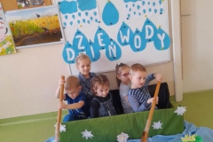 Pięć dzieci siedzi w Zielonej łódce wykonanej z dużego pudełka, dwoje z nich naśladuje wiosłowanie, za dziećmi tablica z napisem dzien wody. Przed nimi na niebieskim materiale ułożone lilie wodne, papierowe łódki, żabki.