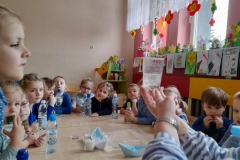 Dzieci siedzą przy stolikach obserwują co dzieje się z rodzynkami wrzuconymi do szklanki z wodą gazowaną. Na stole widać inne przedmioty potrzebne do doświadczeń z wodą np. papierowe łódki, sól, cukier, woda gazowana w butelce.