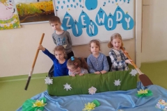 W Zielonej łódce wykonanej z pudełka siedzi 5 dzieci, 2 chłopców trzyma w reku wiosła, za nimi  tablica z napisem dzień wody. Przed nimi rozłożony niebieski materiał na którym ułożone są lilie wodne, zielone żabki oraz papierowe łódki.