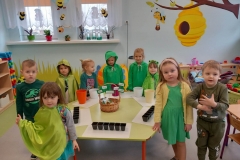 .Sala przedszkolna . Grupa dzieci ubrana w  stroje w odcieniach koloru zielonego zgromadzona wokół sześciokątnego stolika na którym stoją doniczki i pojemniki z ziemią . Na środku stolika koszyk , a w nim torebki z nasionami różnych roślin . W koszyku umieszczone są tabliczki z nazwami roślin.