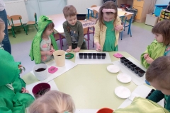 Przy stoliku grupa dzieci ubrana na zielono . Dzieci wkładają do doniczek lub pojemników nasiona wybranych roslin.