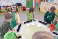 Przy stoliku grupa dzieci ubrana na zielono . Dzieci wkładają do doniczek lub pojemników nasiona wybranych roslin.