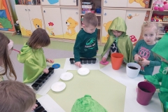 Przy stoliku grupa dzieci ubrana na zielono . Dzieci wkładają do doniczek lub pojemników nasiona wybranych roslin.