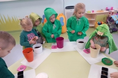 Przy stoliku grupa dzieci ubrana na zielono . Dzieci wkładają do doniczek lub pojemników nasiona wybranych roslin.