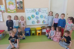 Zdjęcie wykonane w sali przedszkolnej. Siedmioro dzieci stoi w półkolu, tyle samo siedzi przed nimi na podłodze.  W tle na białej tablicy jest napis oraz prace plastyczne dzieci w kształcie kół.