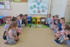 Dzieci siedzą na podłodze w dwóch rzędach. Pośrodku stoją cztery kosze na śmieci  w różnych kolorach. Przed koszami leżą różne śmieci.