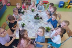 Grupa dzieci siedzi przy stoliku. Przed nimi doniczka z posadzonymi kwiatami.