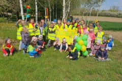 Zdjęcie przedstawia dzieci ubrane w żółte kamizelki odblaskowe. Część z nich kuca, pozostałe stoją. Trzymają w rękach kwiaty oraz balony.