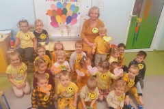 Grupa dzieci siedzi na podłodze. Dwóch chłopców siedzi na ławeczce. Ubrane w żółte stroje. W tle biała tablica, a na niej żółte słoneczko oraz kolorowe paski papieru.