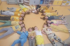 Dzieci leża na podłodze tworząc promienie słońca. W tle szafka z szufladkami.