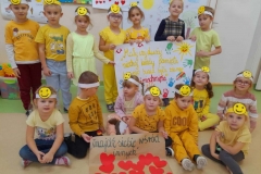 Grupa dzieci ubrana na żółto, część z nich  która stoi w tyle trzyma w rękach plakat z kolorowym napisem i odbitymi rączkami. Kilkoro dzieci siedzi na podłodze, przed nimi drzewko życzliwości wykonane z czerwonych serduszek.