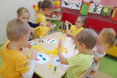 Grupa dzieci siedzi przy stoliku, naklejają oczy  buźki i kolorowe włosy na białe kółko tak by powstała uśmiechnięta bużka.