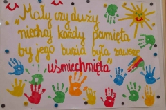 Na dużym arkuszu papieru napis: Mały czy duży niech każdy pamięta, by jego buzia była zwsze uśmiechnięta. Wokół napisu odbite kolorowe rączki dzieci.
