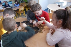 Dzieci w kolorowych bluzach siedzą przy stoliku przedszkolnym. Segregują leżące na stole żółte monety do plastikowych, ciemnych pojemników. Dziewczynka z prawej strony ma kucyk. Ubrana jest w różową bluzkę. Siedzący z lewej strony chłopiec, odwrócony tyłem, ubrany jest w szarą bluzę z kapturem. Chłopiec siedzący na wprost ma czerwoną bluzę. Wybiera monety z dłoni. Obok niego siedzi chłopiec w szarej bluzie. Zbiera monety ze stołu. Chłopca w żółtej bluzie widać tylko częściowo. Za nimi widoczny jest stolik z trójką rysujących dzieci i szafki przedszkolne.