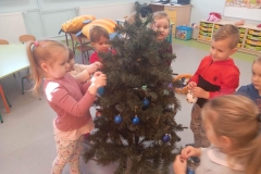 Sala przedszkolna . Grupa dzieci ubrana na kolorowo stoi wokół choinki i zawiesza na niej bombki w odcieniach koloru niebieskiego