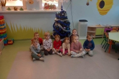 Dzieci w sali przedszkolnej siedzą przed choinką ubraną w bombki, łańcuchy w kolorach niebieskich i srebrnych. W tle ściana z częścią okien , na jednym dekoracja Mikołaja na sankach. Z prawej strony szafki kącika kuchennego i część stolika dla dzieci.