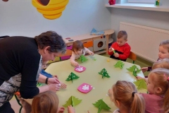 Dzieci siedzą przy stoliku . Na stoliku przestrzenne papierowe zielone choinki i na dwóch różowych talerzykach białe śnieżynki. Każde dziecko nakleja śnieżynki na swoją papierową choineczkę.