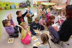 Kilkoro  dzieci siedzi  na podłodze,  pozostałe  stoją tworząc półkole.  Pośrodku na podłodze leży żółty  karton z kilkoma emotkami. W tle  jasne szafki.