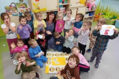 Kilkoro  dzieci siedzi na podłodze, za nimi siedzą pozostałe dzieci, obok pani. W tle dekoracja z kolorowymi emotkami.