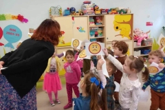 Dzieci ubrane w kolorowe stroje stoją  w rozsypce, nad nimi pochyla się  pani. W tle jasne szafki.