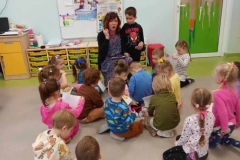 Dzieci siedzą na podłodze. Przed  nimi na krzesełku siedzi pani.