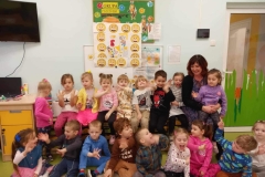 Dzieci siedzą na podłodze,  przed nimi leżą żółte buźki.  Przed nimi na krzesełku siedzi pani.