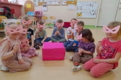 Dzieci siedzą w półkolu, na nosie założone różowe okulary, po środku nich różowe pudełko.