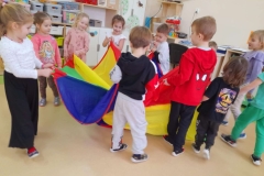 Dziecko siedzi na środku chusty a imacyjnej, reszta dzieci krę i chustą dookoła.