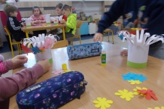 Przy stoliku przedszkolnym trzech chłopców wykonuje przestrzenną pracę plastyczną. Na sklejonych z kartki rulonach z naciętymi paskami u góry  naklejają kolorowe kwiaty wycięte z papieru. W głębi widoczne dwa stoliki, przy których również wykonywane są te działania dzieci.