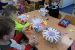 Przy stoliku przedszkolnym siedzi trzech chłopców i jedna dziewczynka. Kolorują one obrazki konturowe z motywem pisanki. Na stoliku leża piórniki i pojemniki  z kredkami.