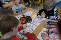 W sali przedszkolnej przy dwóch stolikach siedzą dzieci. Przy pierwszym stoliku widoczne są tylko ich ręce, przy drugim w głębi sali widać czworo dzieci. W rogu po lewej stronie widać częściowo postać kobiety. Na stolikach leżą piórniki, kleje oraz wycięte z papieru kolorowe kwiaty. Przed dziećmi stoją szerokie rulony z białego papieru z naciętymi u góry i wywiniętymi paskami. Do nich dzieci przyklejają kwiaty.