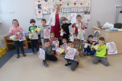 Dzieci przedszkolne pozują na tle ściany. Te z tyłu stoją, miedzy nimi w środku znajduje się kobieta w czerwonej sukience i białym żakiecie. Dzieci z przodu siedzą na podłodze. Wszystkie trzymają w rękach swoje prace - koszyki z kwiatami wykonane    z papieru oraz obrazki z pisankami. Na ścianie za nimi widoczna jest tablica z literami   i cyframi.